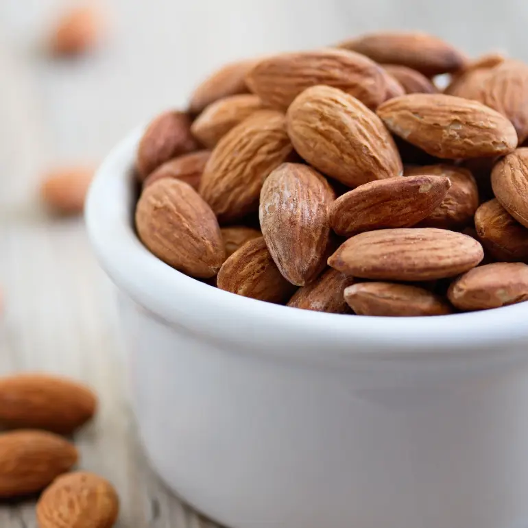 A close-up of a white bowl filled with whole almonds, placed on a wooden surface. Several almonds are scattered around the bowl.