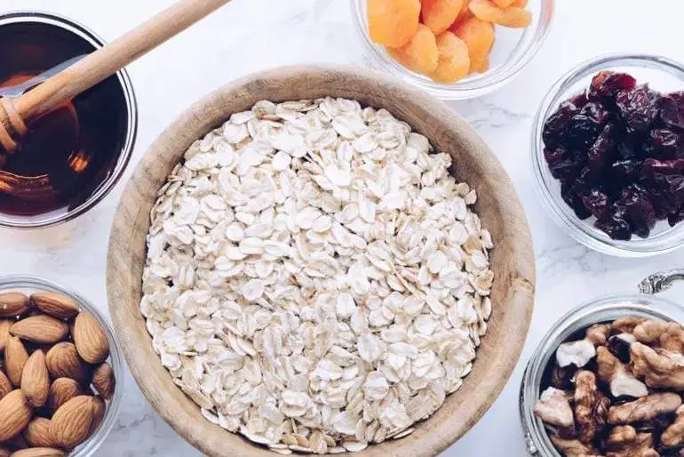 A wooden bowl filled with rolled oats surrounded by small glass bowls containing honey with a dipper, dried apricots, dried cranberries, almonds, and walnuts.